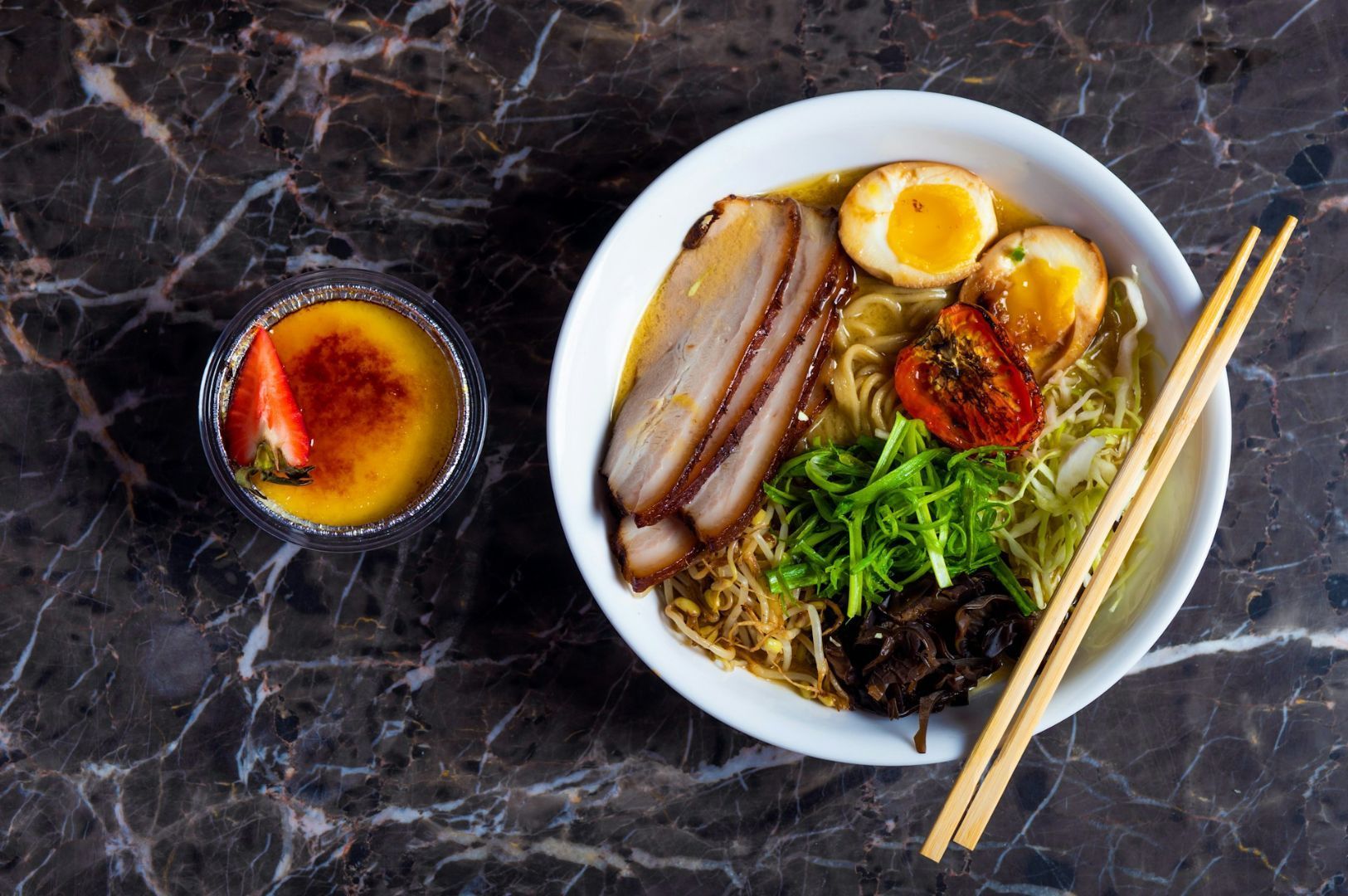 A bowl of ramen with sliced pork, a halved soft-boiled egg, scallions, and mushrooms on a marble surface. Chopsticks rest on the bowl. A small crème brûlée with a strawberry is beside it. The scene conveys a sense of warmth and culinary delight.