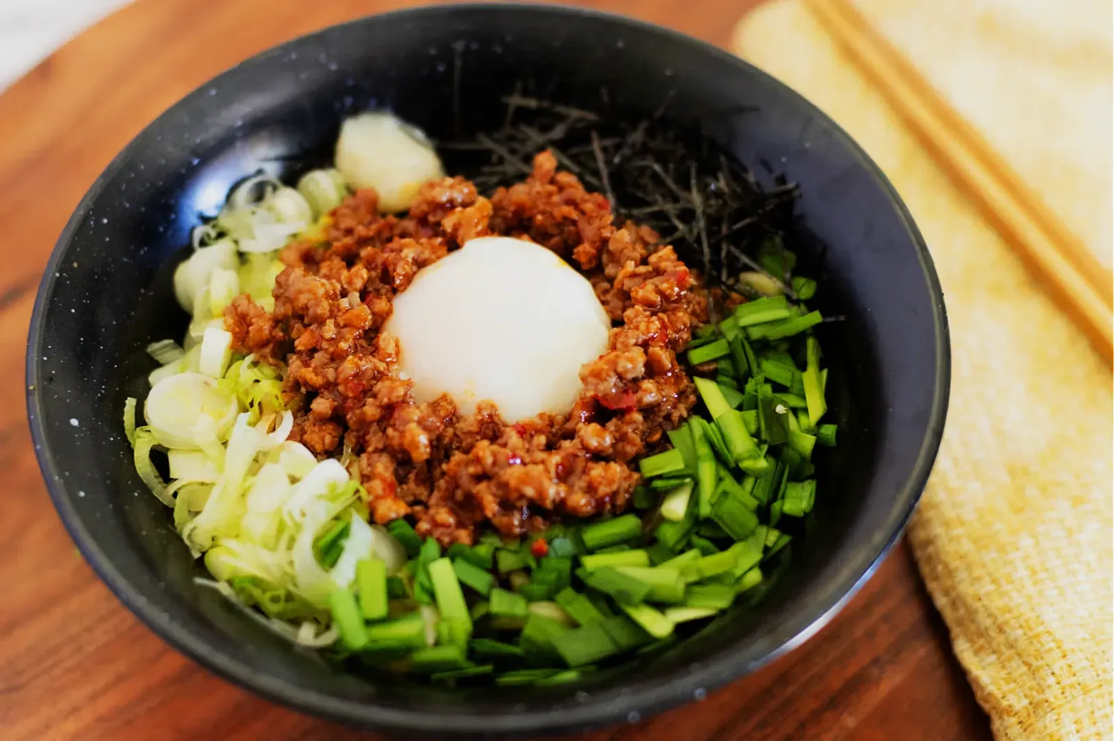 A black bowl with a soft-boiled egg nestled in minced meat, surrounded by sliced scallions, green veggies, and seaweed. Chopsticks rest on a yellow napkin.