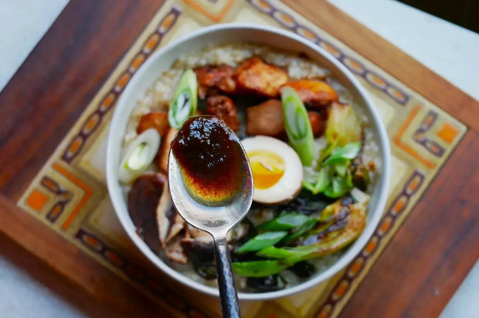 A close-up of a spoon with dark sauce above a bowl of ramen topped with boiled egg, scallions, grilled meat, and mushrooms on a patterned placemat.