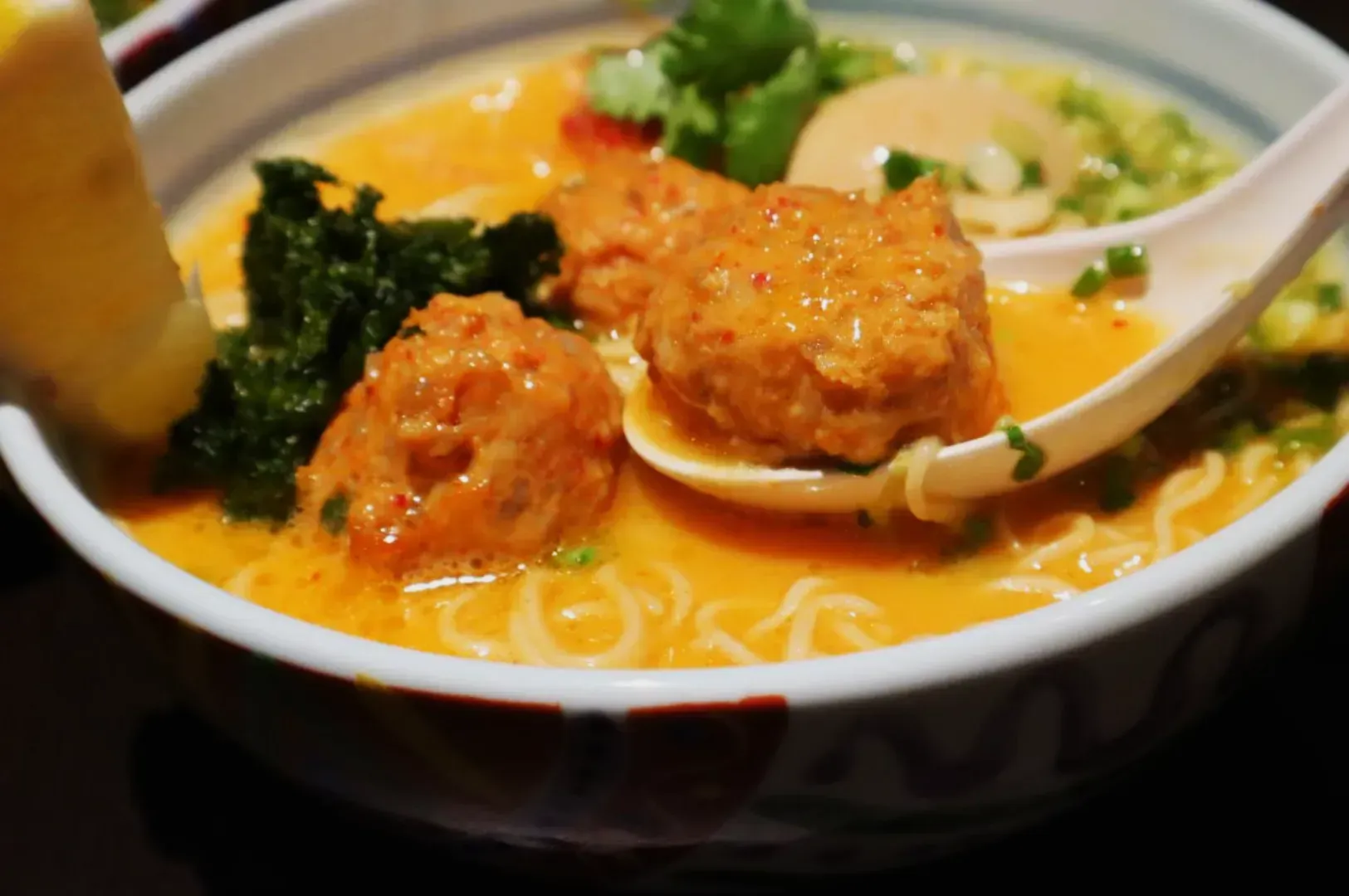 A bowl of ramen featuring vibrant orange broth, meatballs, greens, and a halved egg. A lemon wedge is on the rim, and a spoon holds a meatball.