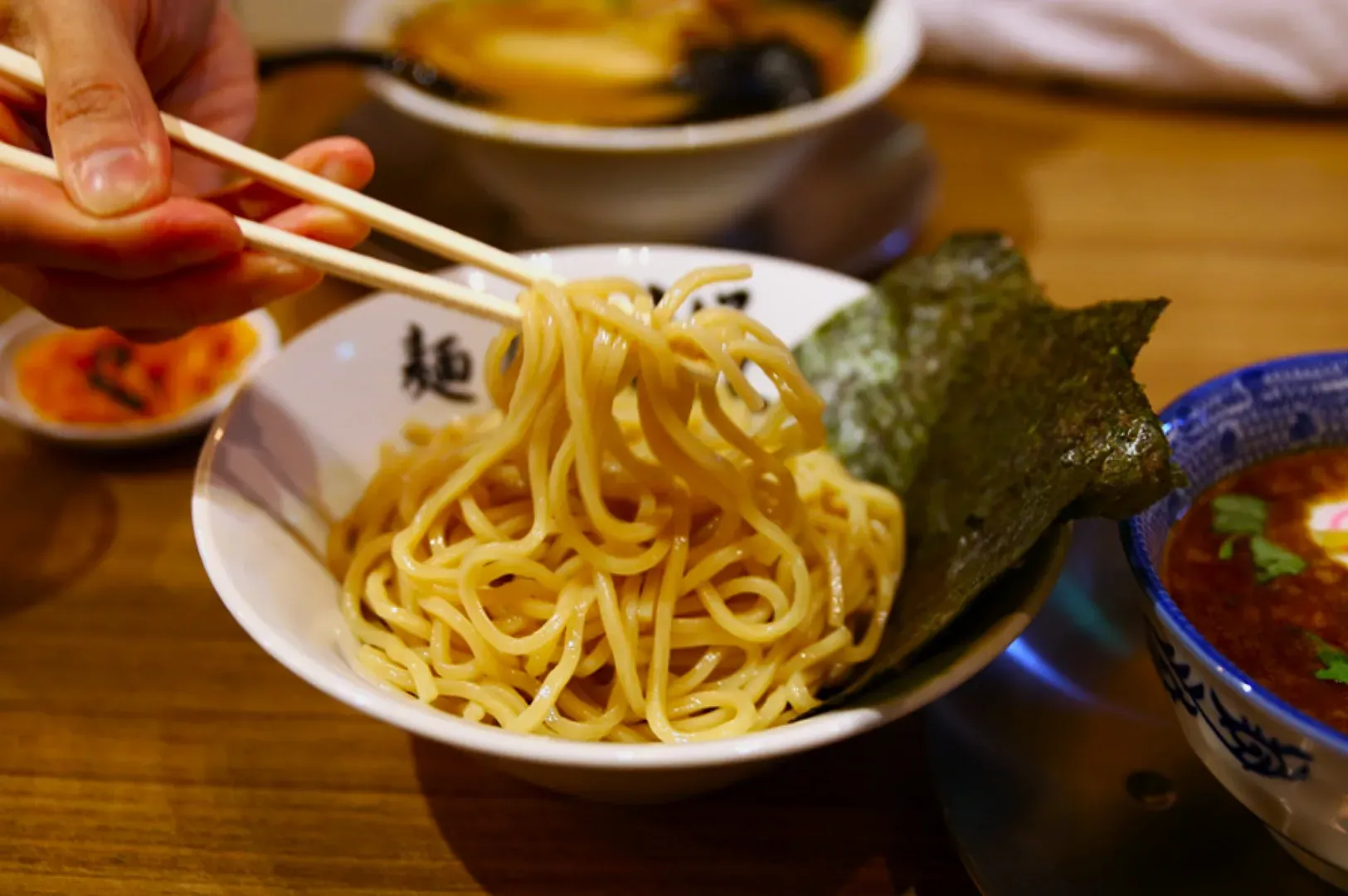 A hand with chopsticks lifts noodles from a white bowl, accompanied by a sheet of nori. A side dish and soup bowl sit on the wooden table, creating a cozy dining scene.