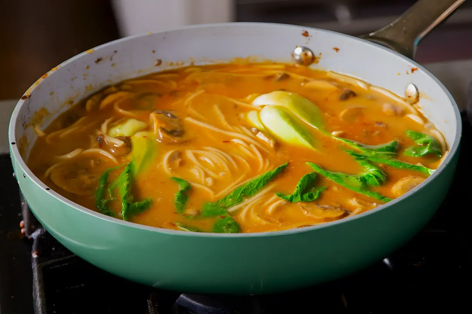 A green pan on the stove holds a simmering curry noodle soup with bok choy, mushrooms, and spaghetti, creating a warm and inviting meal.