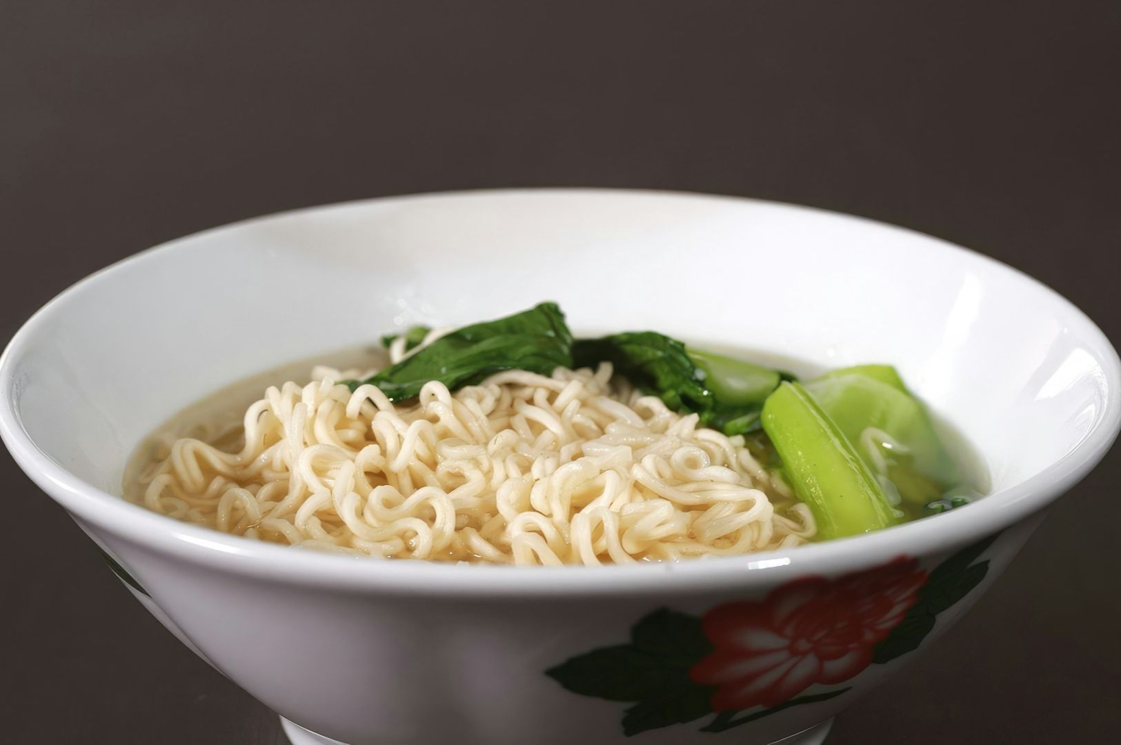 A white bowl filled with cooked instant noodles and vibrant green bok choy, placed on a dark background. The bowl has a floral pattern, suggesting a simple, cozy meal.