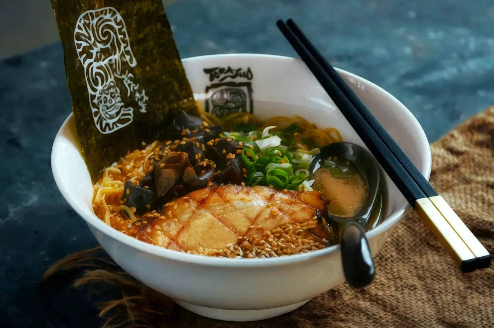 A bowl of ramen with sliced grilled pork, green onions, seaweed, and mushrooms. Chopsticks rest on the bowl, evoking a warm, savory atmosphere.