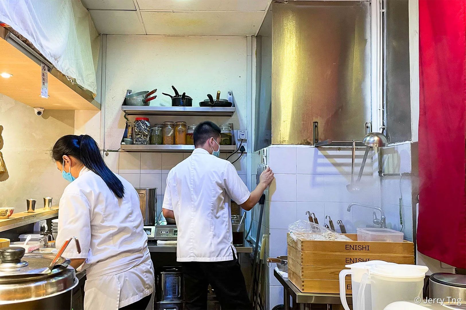 Two chefs wearing white uniforms and masks working in a small, industrial kitchen area, likely preparing ramen or Japanese dishes.