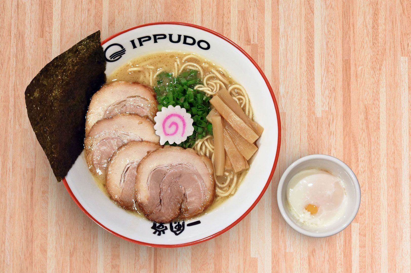 Overhead view of a bowl of Ippudo tonkotsu ramen with chashu pork slices, bamboo shoots, nori, scallions, and a slice of narutomaki, served with a soft-boiled egg on the side.