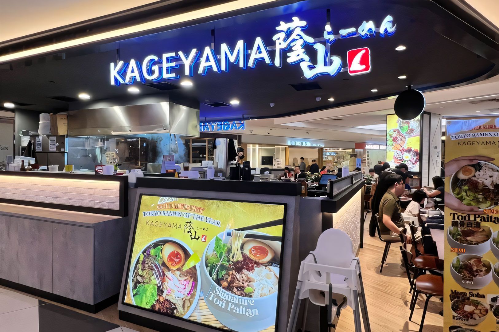 The entrance of Kageyama Ramen restaurant in a mall, featuring a bright blue and white sign, an open kitchen view, and a large display board for their Signature Tori Paitan ramen.