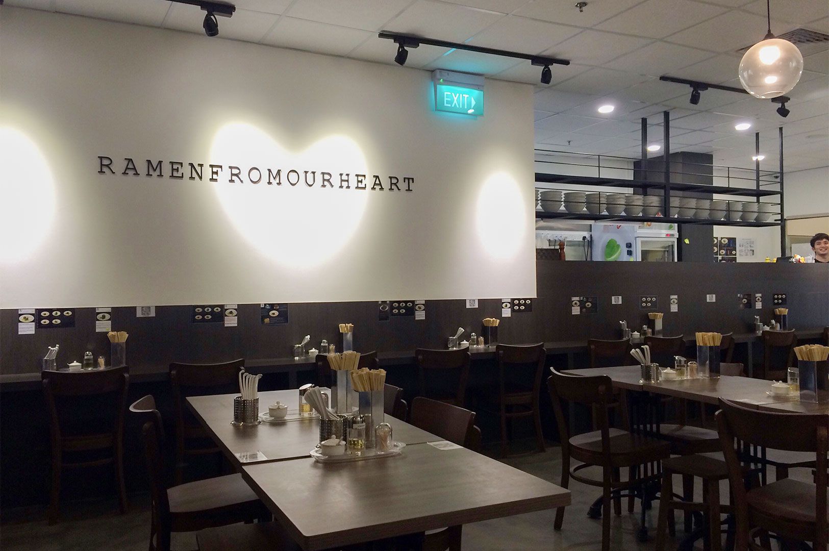 Modern, casual ramen restaurant dining room interior with wooden tables and chairs, featuring a white wall with the text 