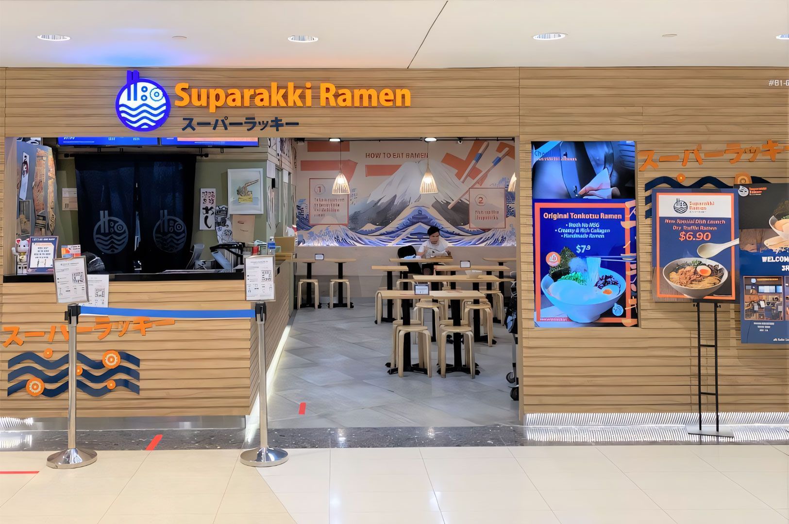 Exterior view of a Suparakki Ramen shop entrance with wood paneling, featuring a dining area mural of Mount Fuji and signage advertising Tonkotsu Ramen deals.