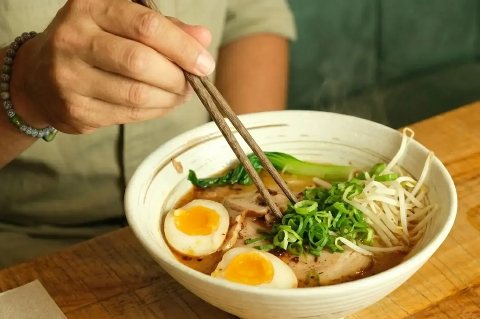 Person using wooden chopsticks to eat a steaming bowl of Kitakata ramen topped with halved soft-boiled eggs, green onions, bean sprouts, bok choy, and pork slices, featuring the signature thick, wavy noodles and clear shoyu broth