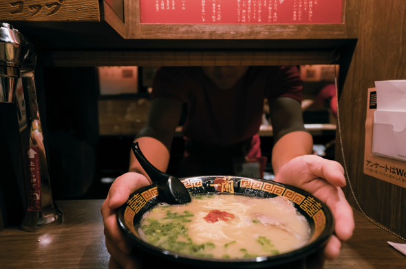 A bowl of rich, umami-packed ramen being discreetly served through a private dining booth window, evoking the minimalist intimacy of Japan’s solo ramen culture. The dish features a deep broth, slices of meat, green onions, and a bold red seasoning, presented in a decorative bowl with a black spoon. The wooden booth, Japanese signage, and napkin dispenser frame the experience, emphasizing quiet focus and culinary reverence—hallmarks of Singapore’s high-end ramen scene inspired by Ichiran-style dining.