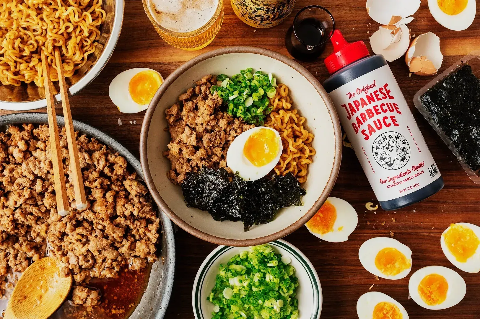 A wooden table with a ramen dish topped with minced meat, green onions, seaweed, and a soft-boiled egg. Surrounding are noodle bowls, barbecue sauce, egg halves, and a beer glass. The scene conveys a rustic and appetizing culinary mood.