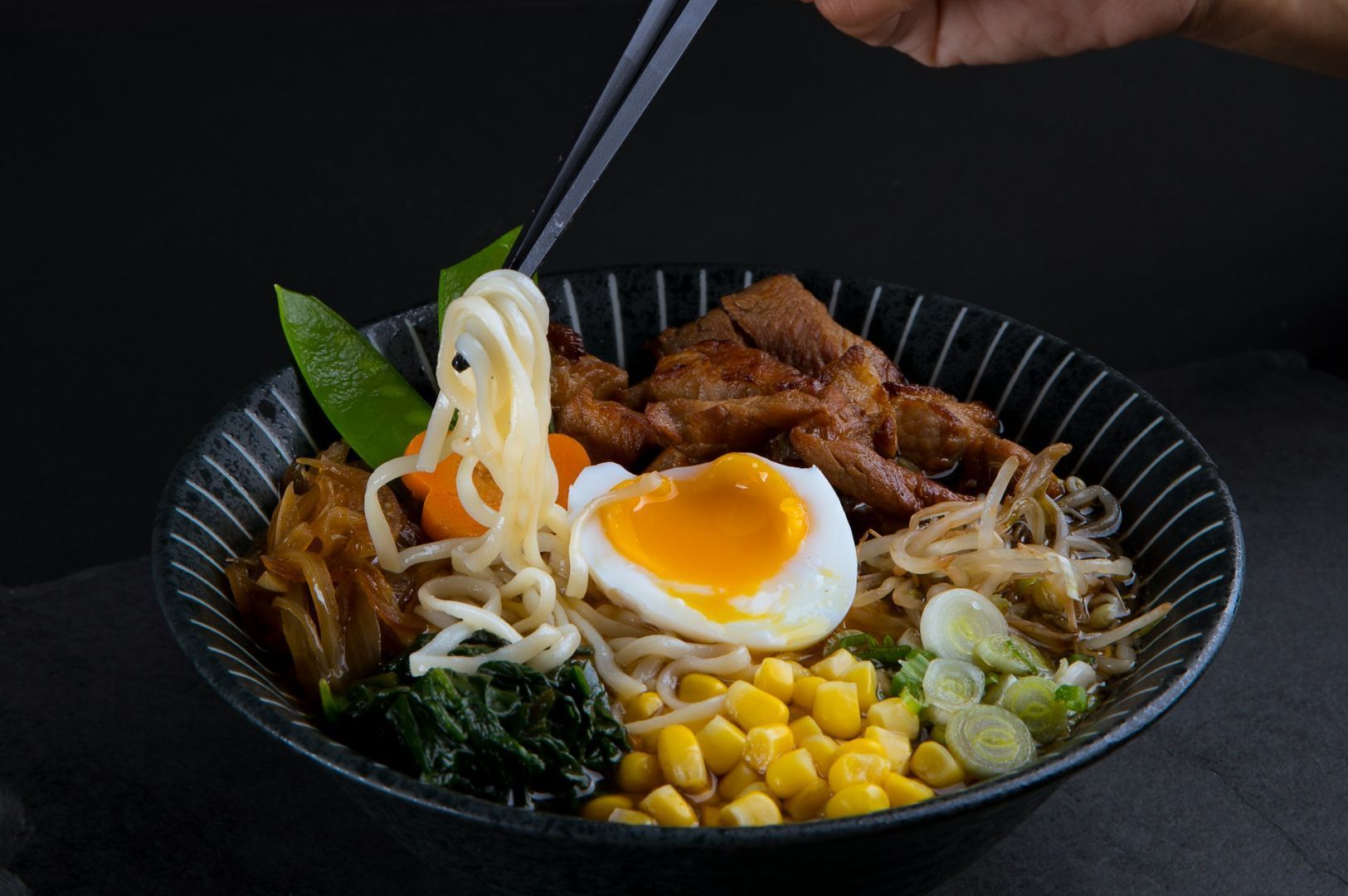 A vibrant bowl of ramen with noodles, a soft-boiled egg, sliced pork, corn, green onions, spinach, carrots, and snow peas, with chopsticks lifting noodles.
