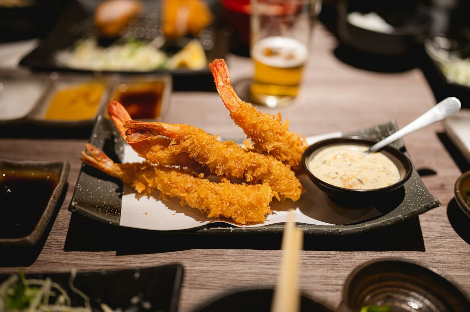 Close-up of three crispy, golden-brown tempura shrimp on a black rectangular plate, served with a creamy dipping sauce. A glass of beer is in the background.