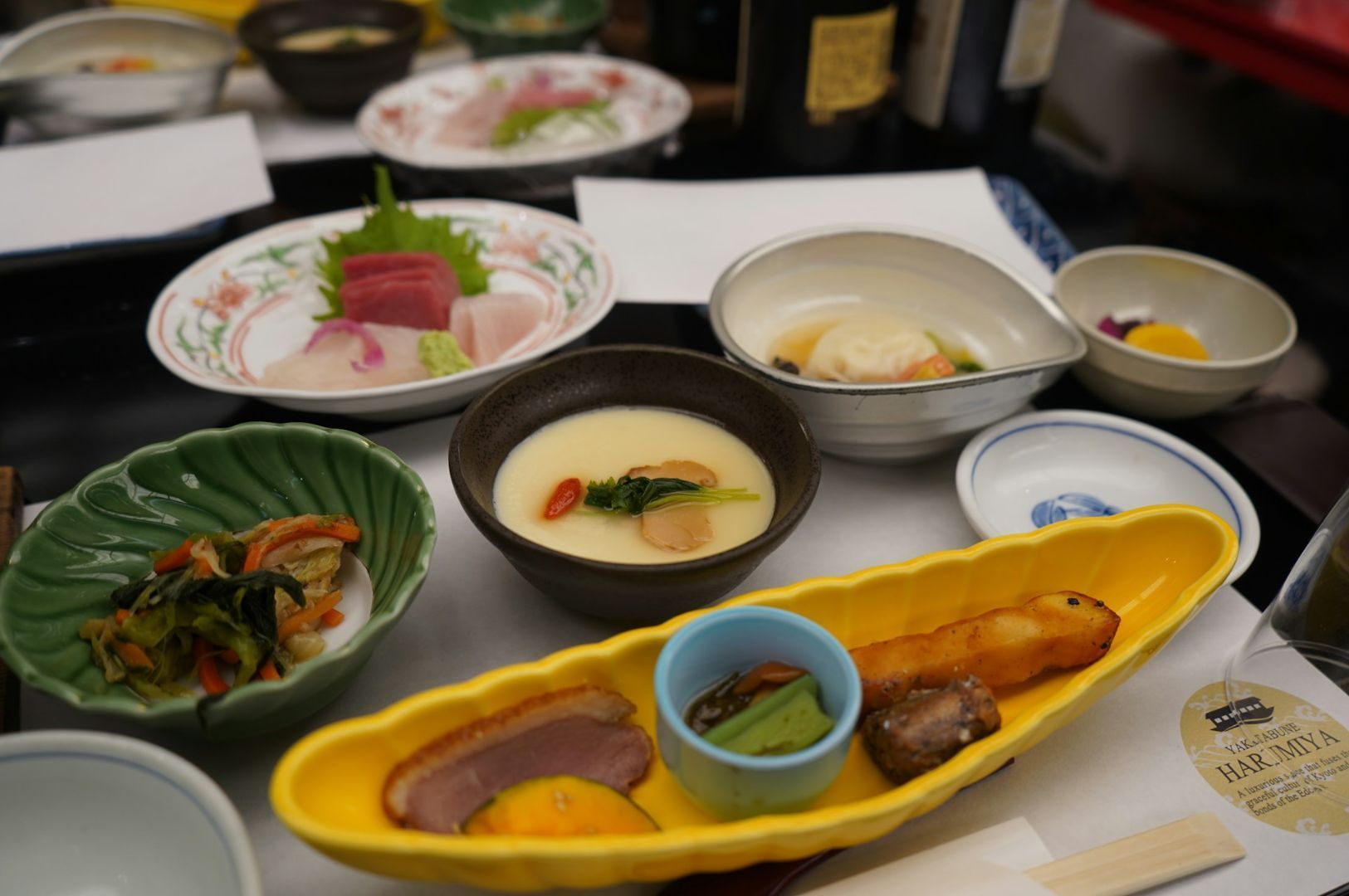 An assortment of Japanese dishes is elegantly arranged, featuring sashimi, steamed custard, colorful vegetables, and grilled fish, conveying a sense of refinement and culinary artistry.