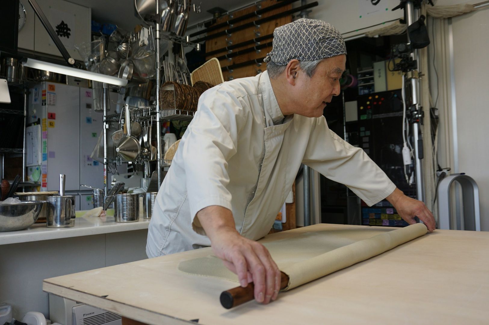 Chef in a white uniform and hair net skillfully rolls dough with a wooden pin in a professional kitchen, surrounded by hanging utensils.