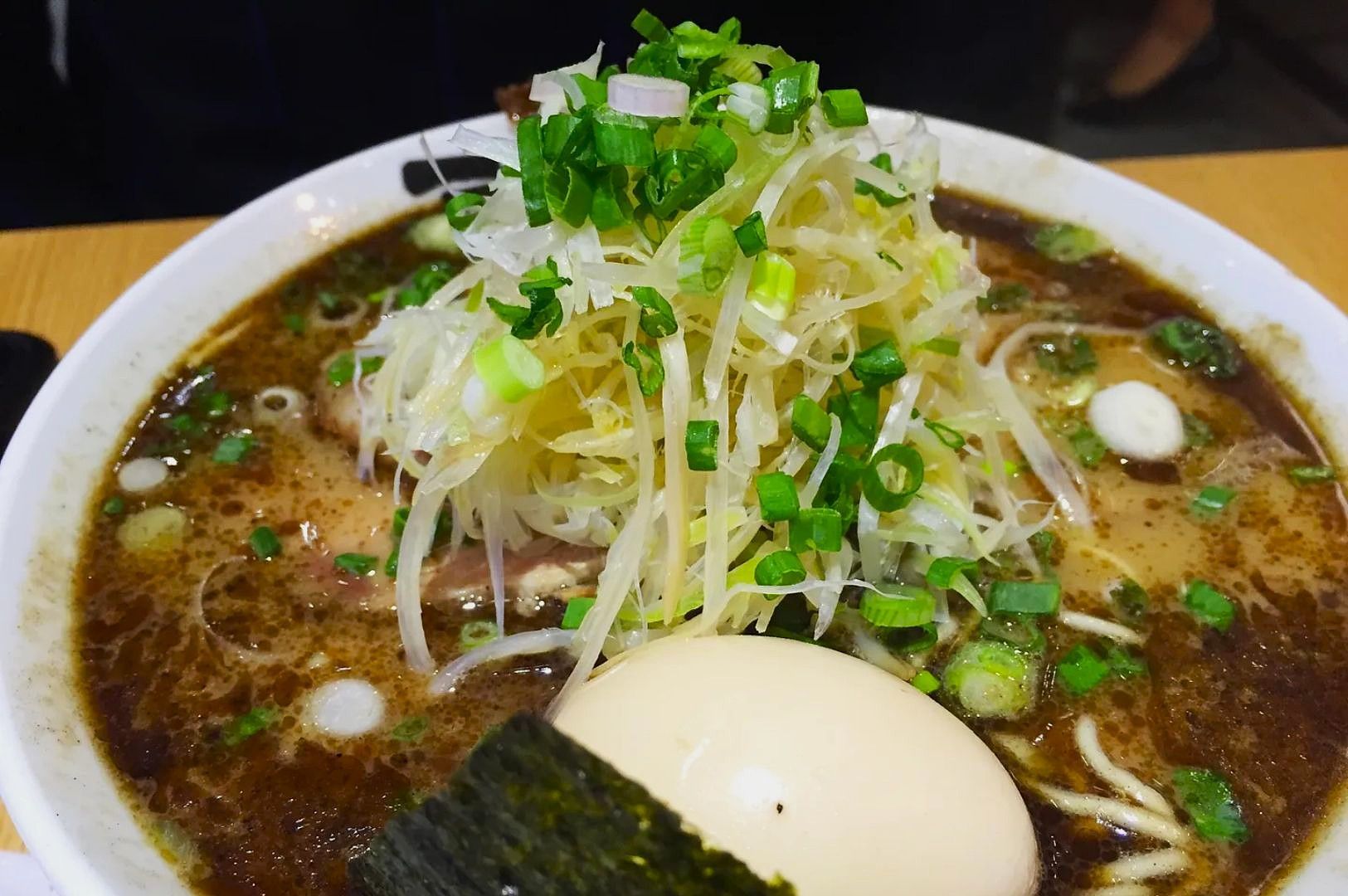 A steaming bowl of dark broth ramen topped with sliced green onions, shredded cabbage, a soft-boiled egg, and a sheet of nori, exuding warmth and savoriness.