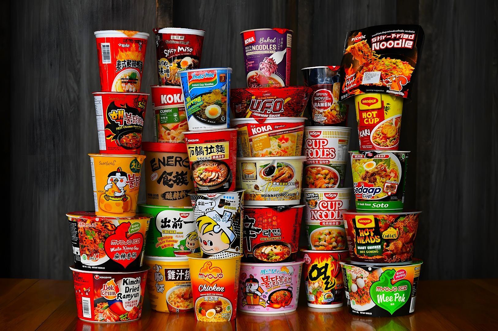 A colorful assortment of instant noodle cups and bowls stacked in a pyramid shape against a dark background. The display features various brands and flavors, exuding a vibrant and diverse culinary selection.
