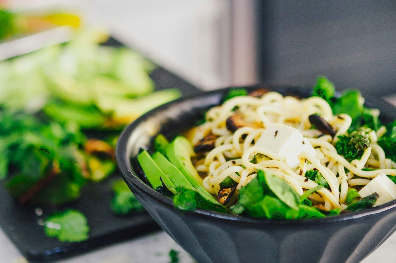 A black bowl filled with stir-fried noodles, tofu, and bright green vegetables like broccoli and avocado slices. The background shows more fresh greens.