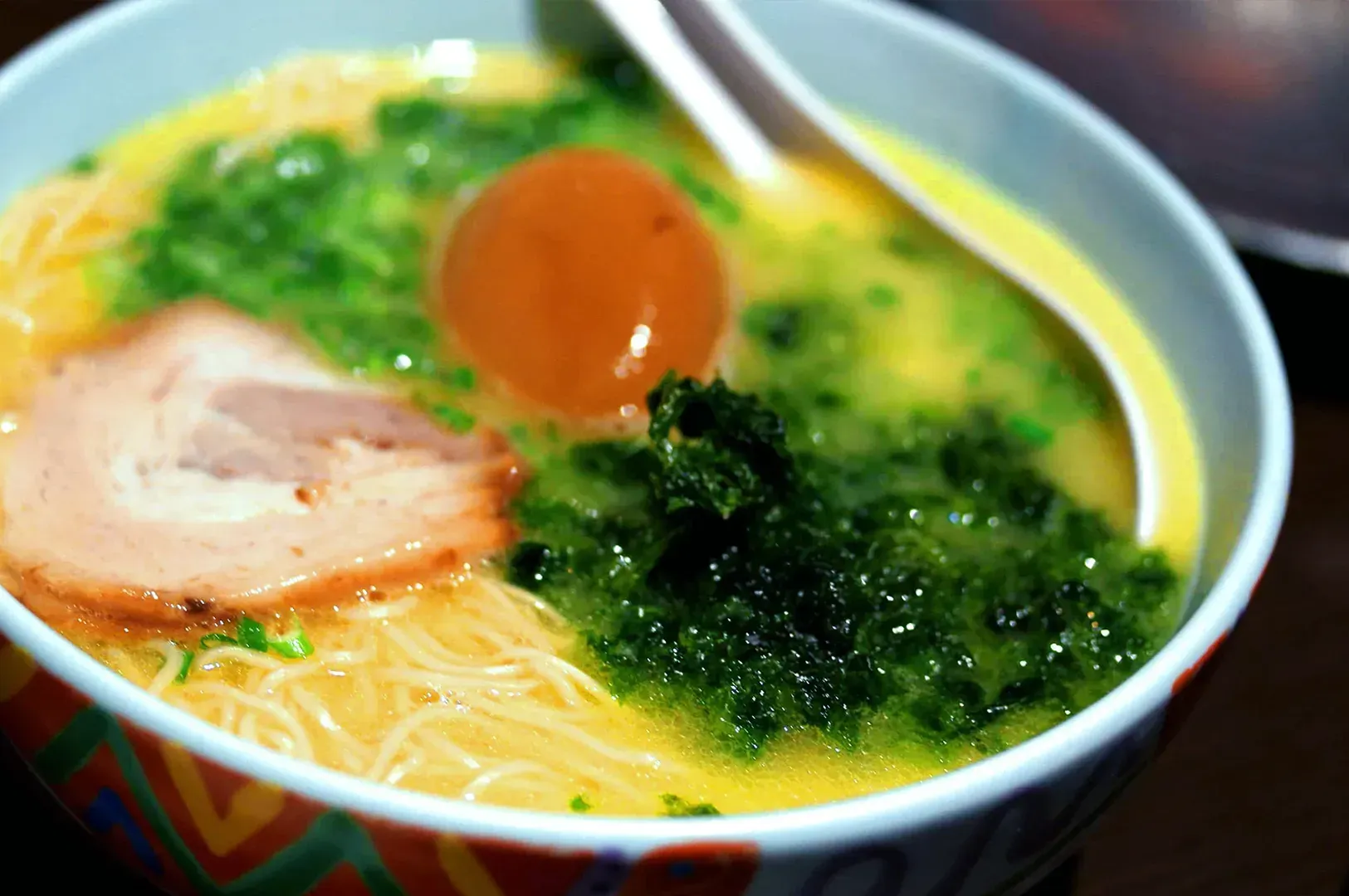 A bowl of ramen with creamy broth, topped with sliced pork, a soft-boiled egg, and green seaweed. A spoon rests on the bowl's edge, evoking warmth.
