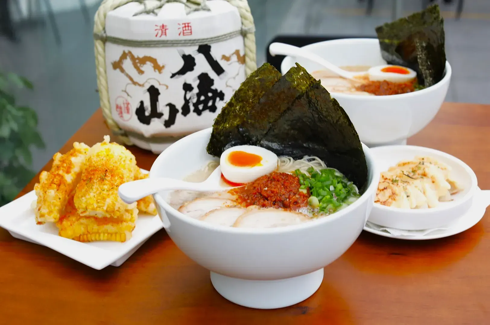 Bowls of ramen with seaweed, half-boiled eggs, green onions, and chili paste. Side dishes include corn tempura and dumplings. A sake barrel is in the background.