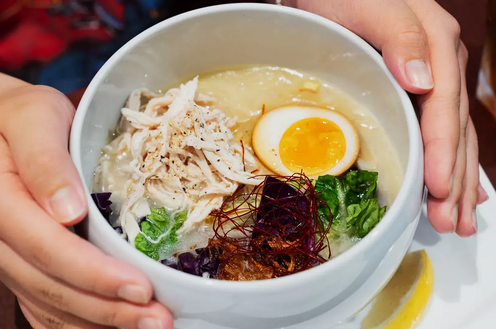 A person is holding a bowl of ramen with shredded chicken, a soft-boiled egg, greens, and red cabbage on top. The presentation looks fresh and inviting.
