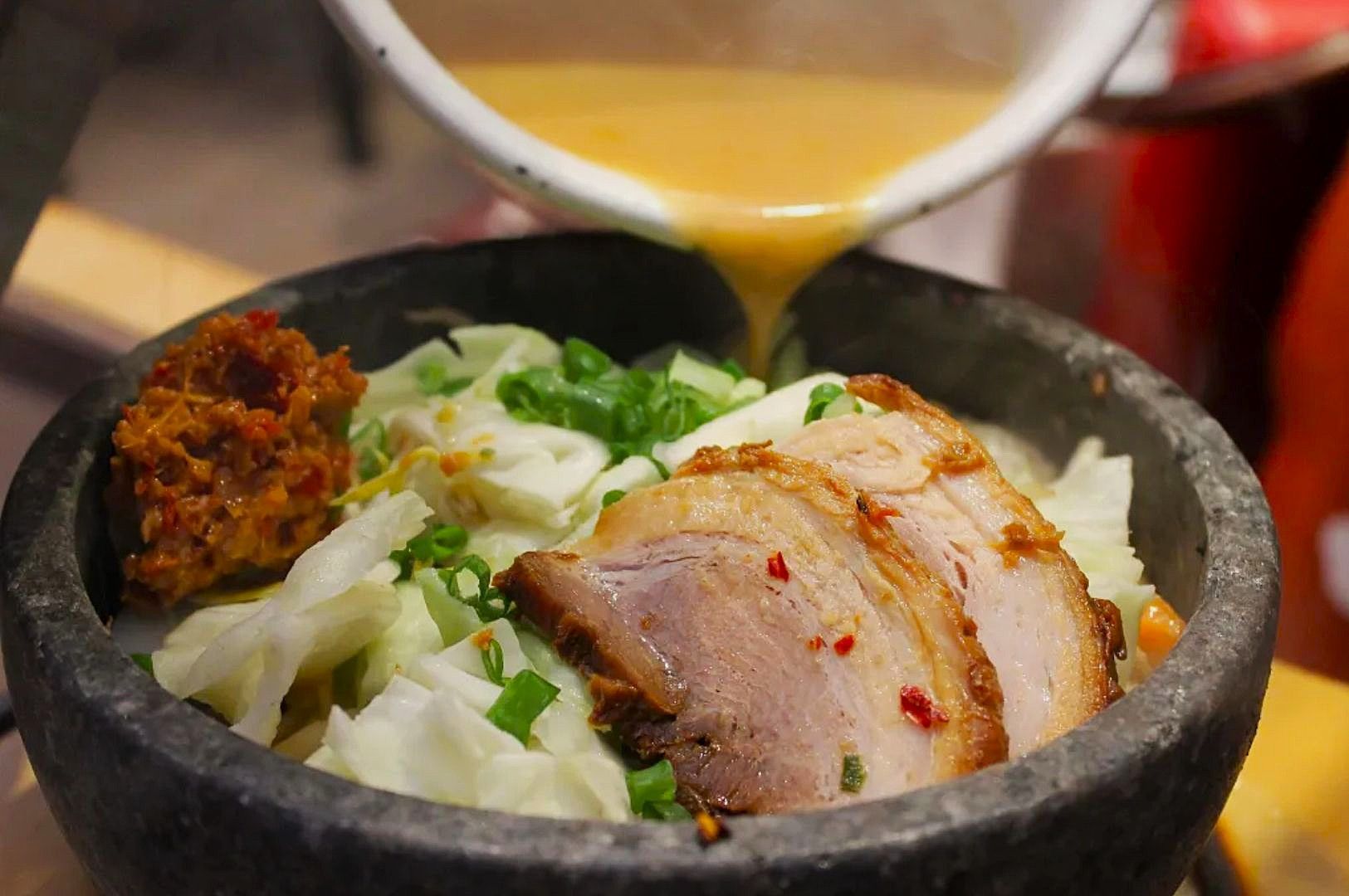 A steaming bowl of ramen with sliced pork, chopped green onions, and cabbage is shown. Rich broth is being poured, enhancing the dish's warmth and flavor.