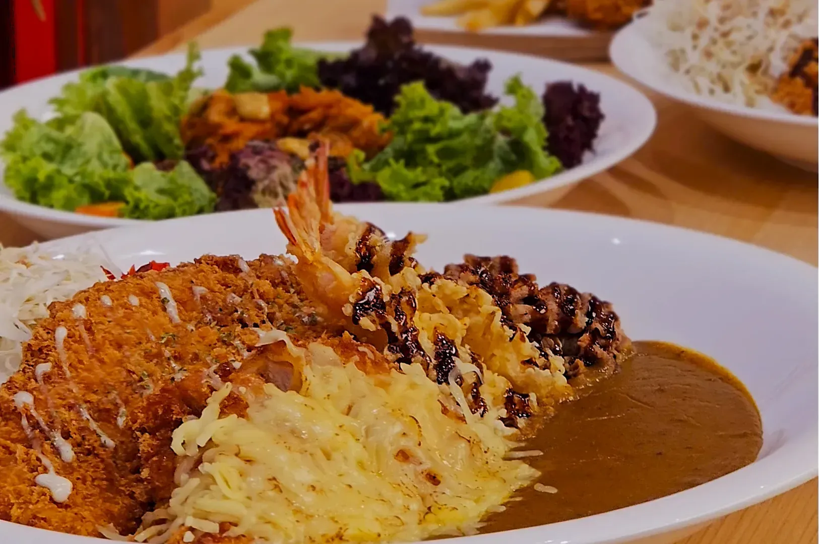 A close-up of a plate with breaded cutlets, shrimp tempura, and sauce on rice. In the background, salad plates with leafy greens are slightly blurred.