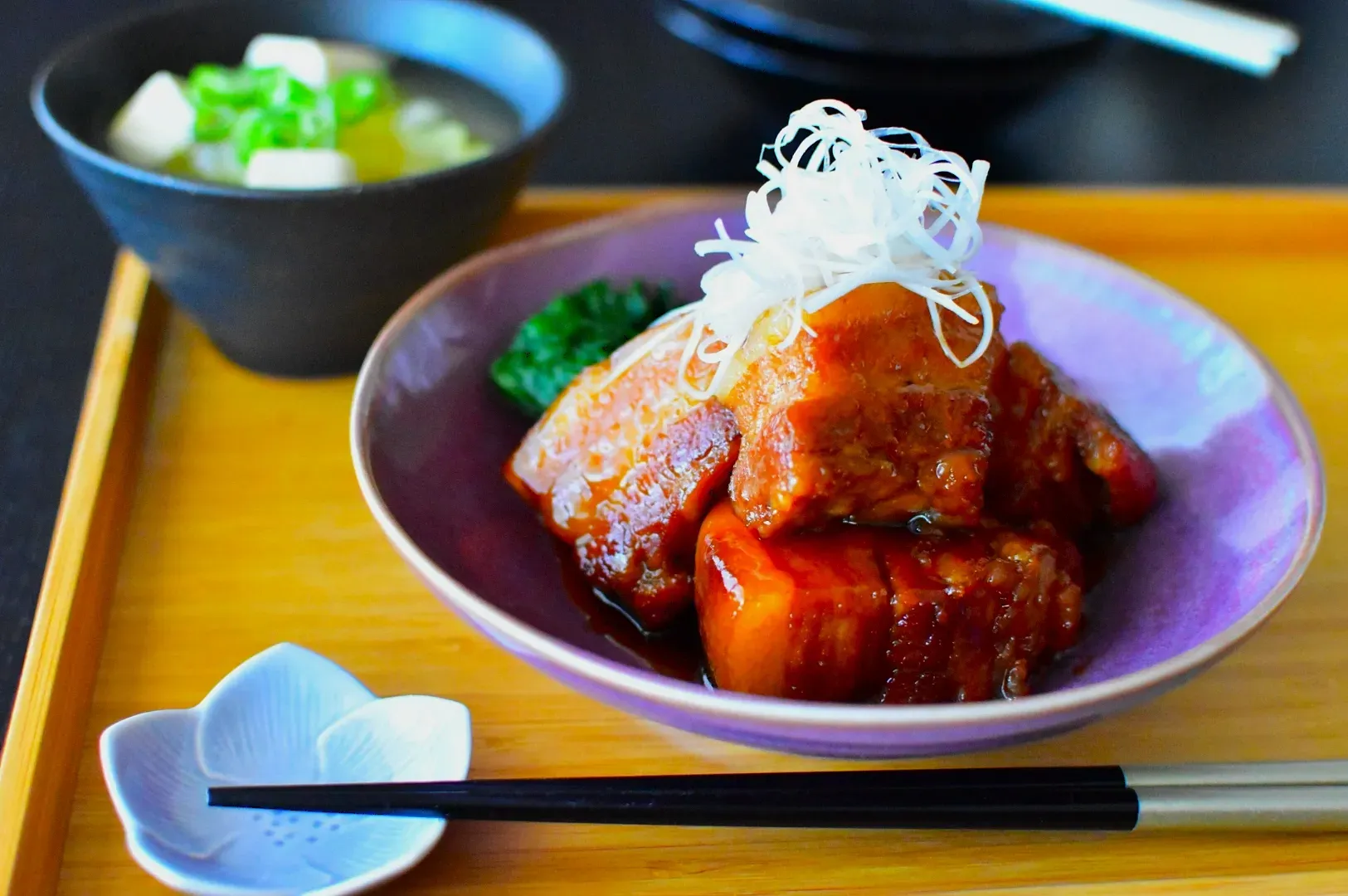 Braised pork belly garnished with shredded white onions in a purple bowl on a wooden tray, accompanied by miso soup and chopsticks, evokes Japanese cuisine.
