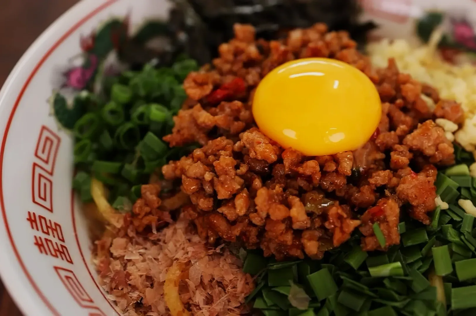 A bowl of ramen topped with minced meat, a raw egg yolk, green onions, bonito flakes, and seaweed. The dish looks vibrant and appetizing.