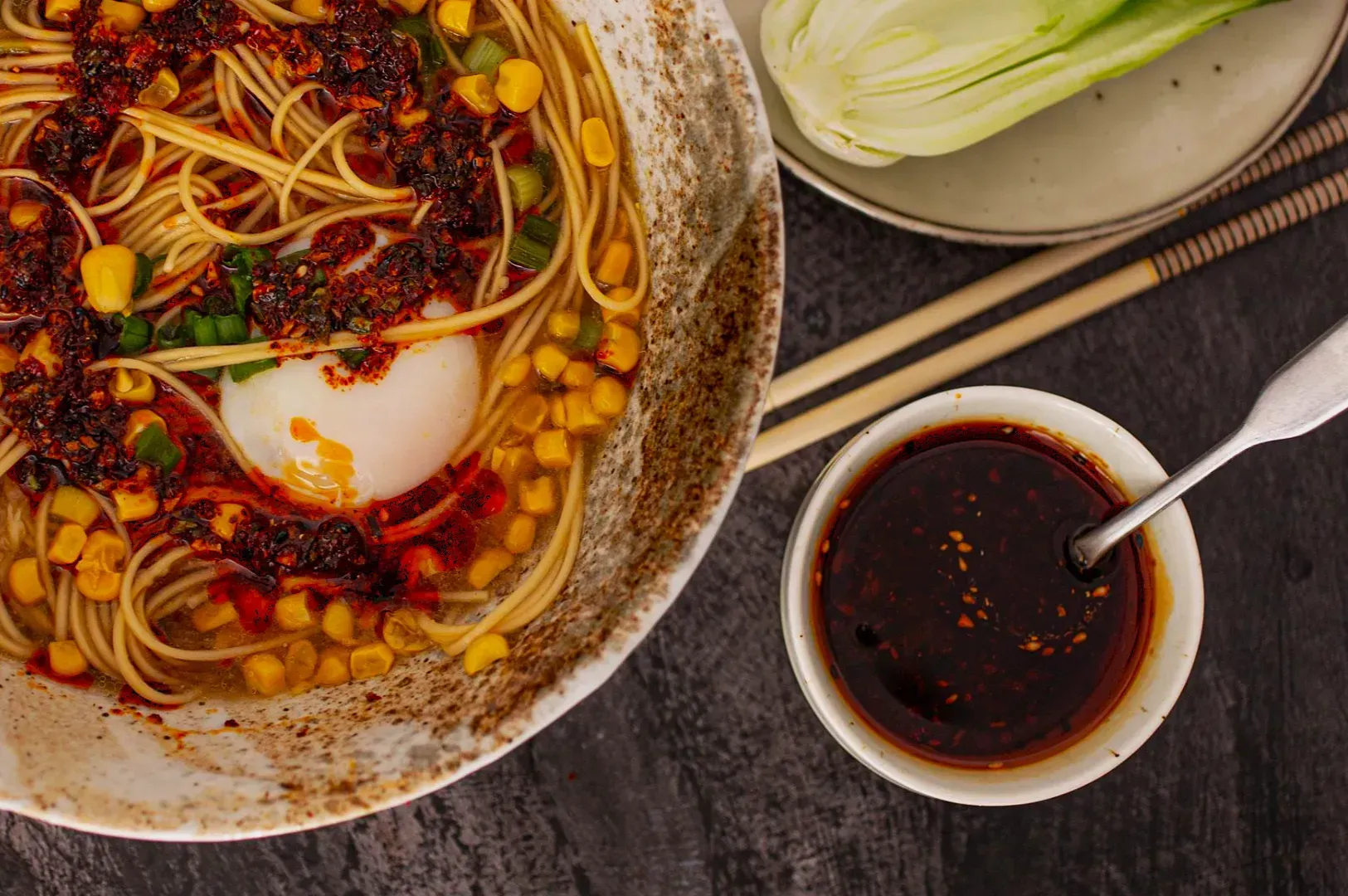 A bowl of ramen with noodles, corn, a soft-boiled egg, and chili oil. Nearby are chopsticks, a sauce dish with more chili oil, and a plate with bok choy.