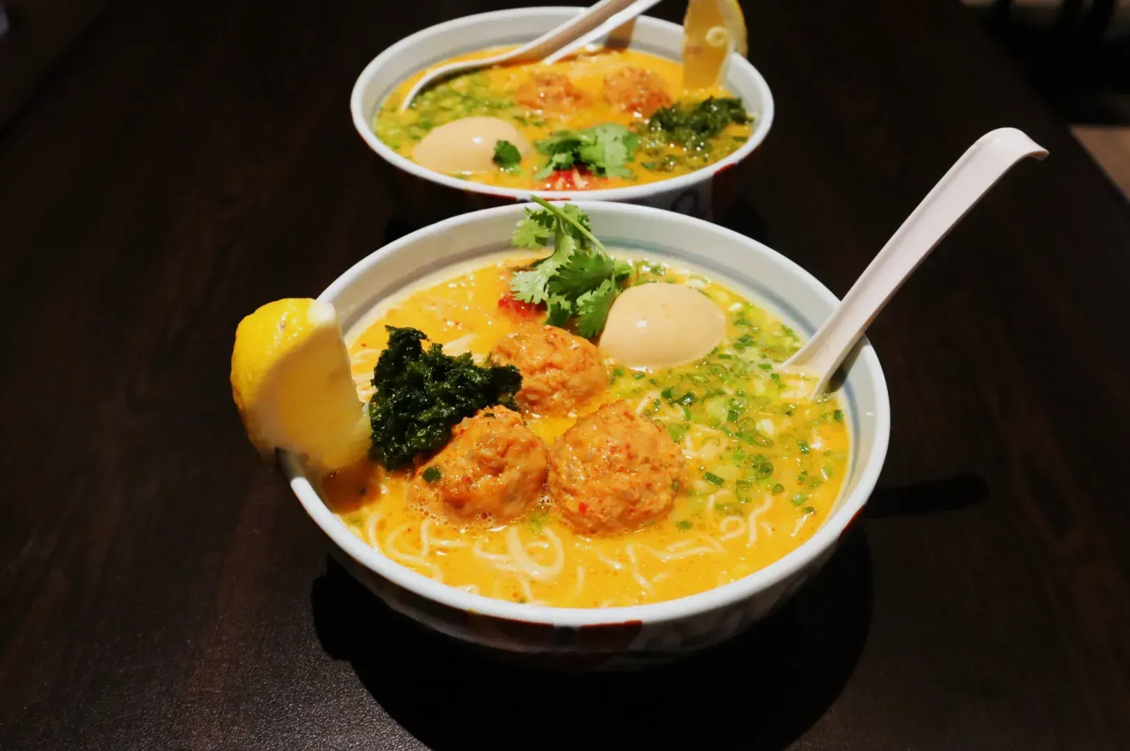 Two bowls of ramen on a dark wooden table. Each has noodles, meatballs, boiled eggs, green herbs, and a lemon wedge. Spoons rest in the bowls.