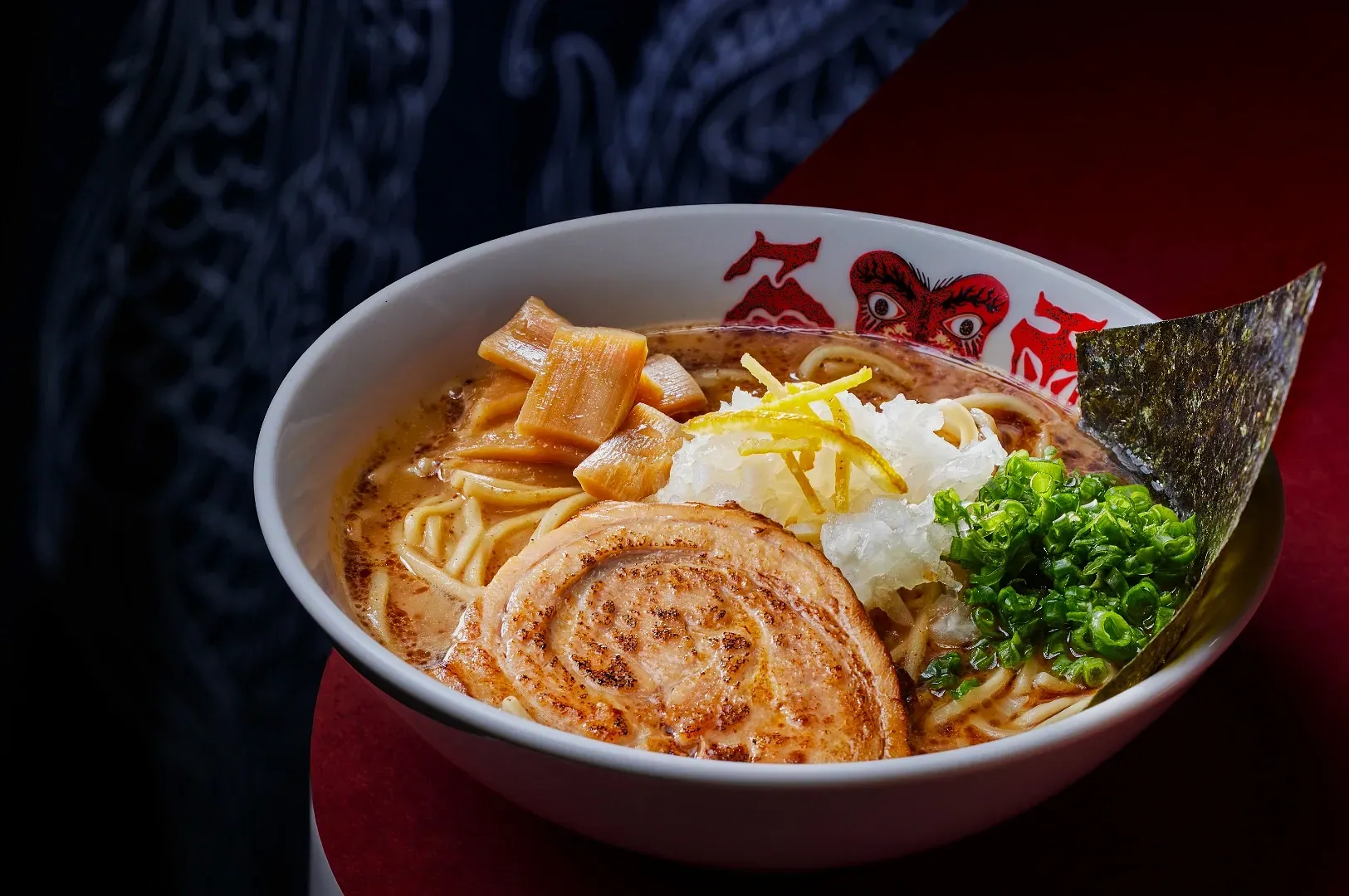 A bowl of ramen with slices of pork, scallions, bamboo shoots, daikon, yuzu zest, and a seaweed sheet, set against a dark, patterned background.