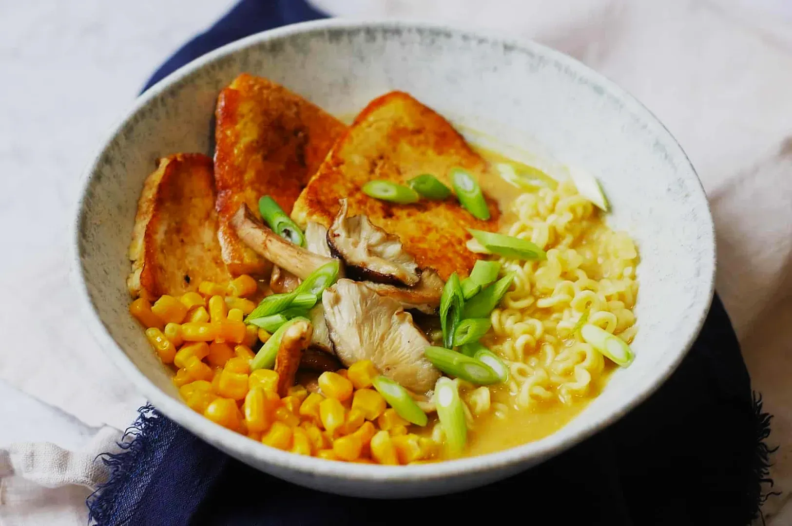 A bowl of ramen with creamy broth, topped with golden tofu slices, mushrooms, sweet corn, and green onions, conveying a warm, savory feel.