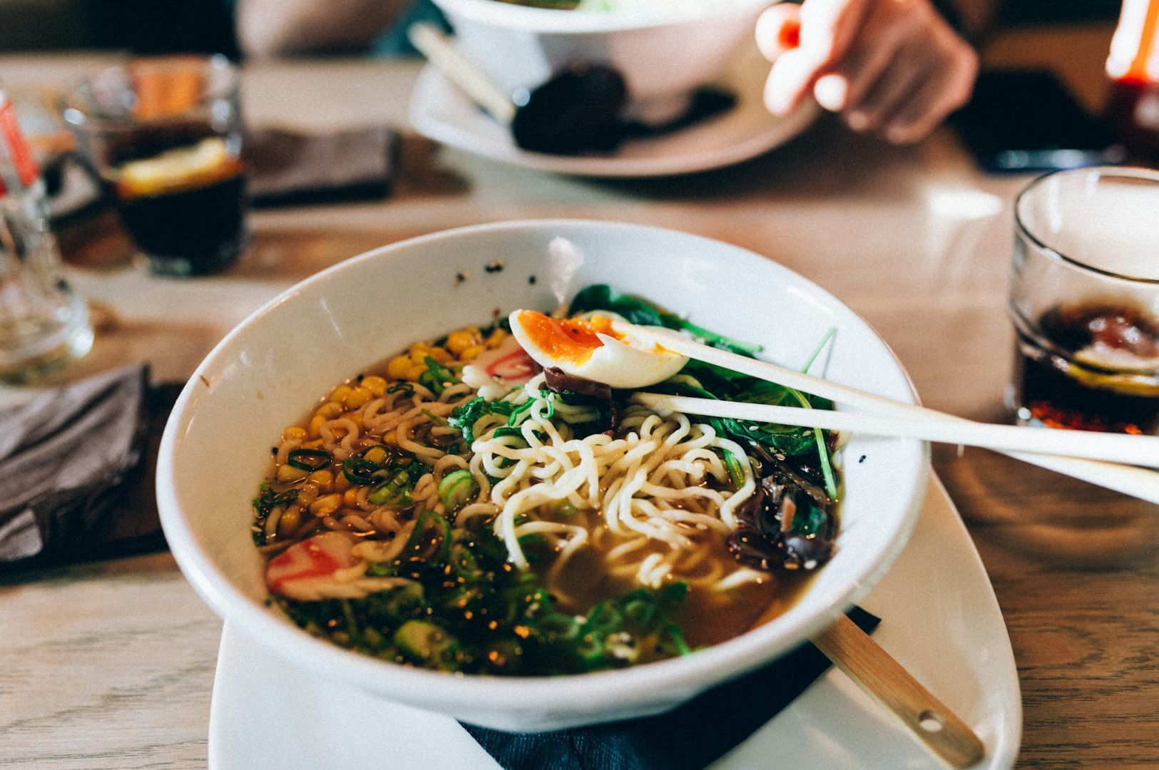 A bowl of ramen with noodles, sliced eggs, corn, seaweed, and greens surrounded by drinks on a wooden table. The setting feels warm and inviting.