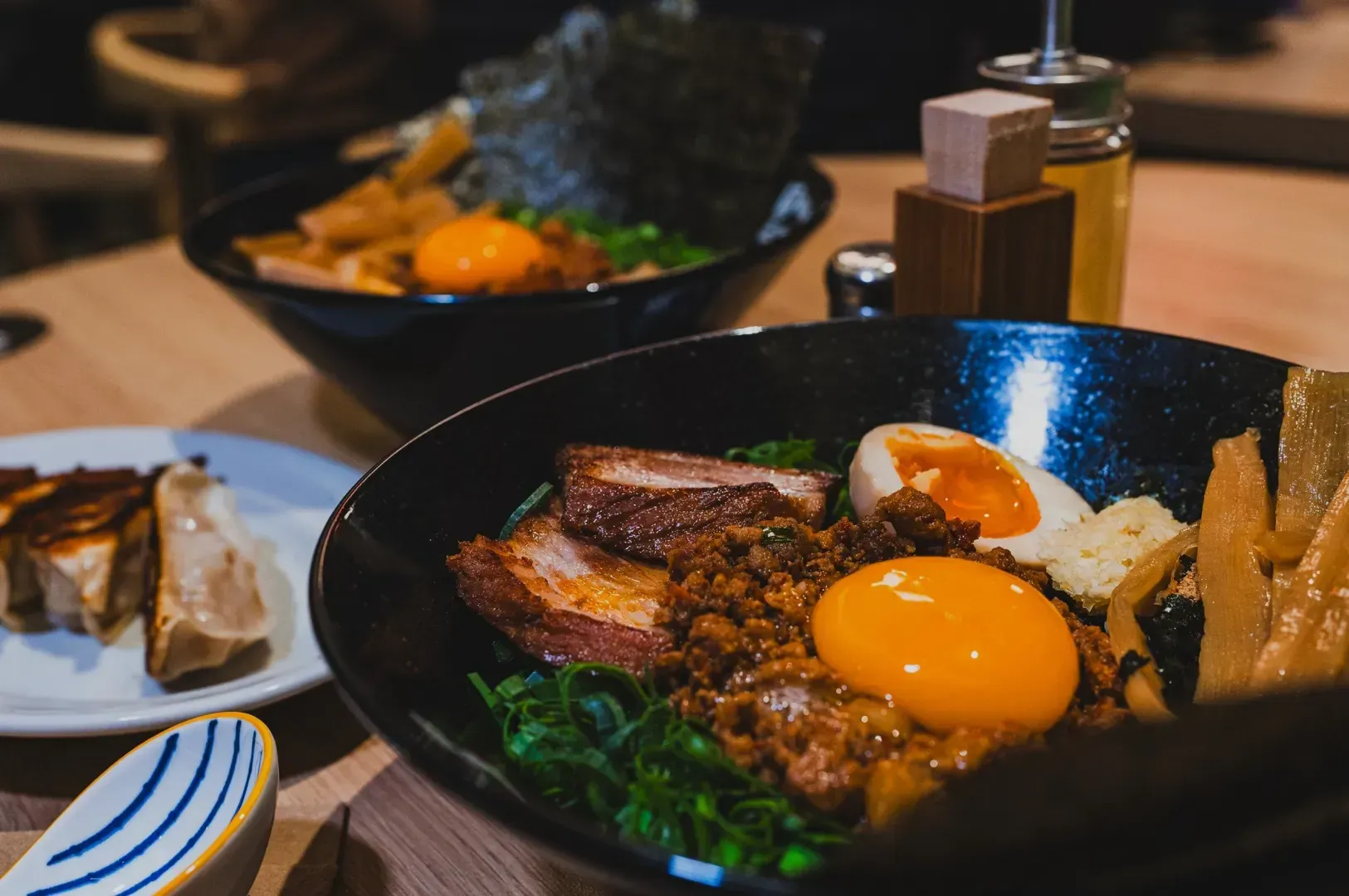 A vibrant bowl of ramen with bright yellow egg yolk, sliced pork, green onions, and seaweed on a wooden table. Gyoza dumplings are visible in the background.