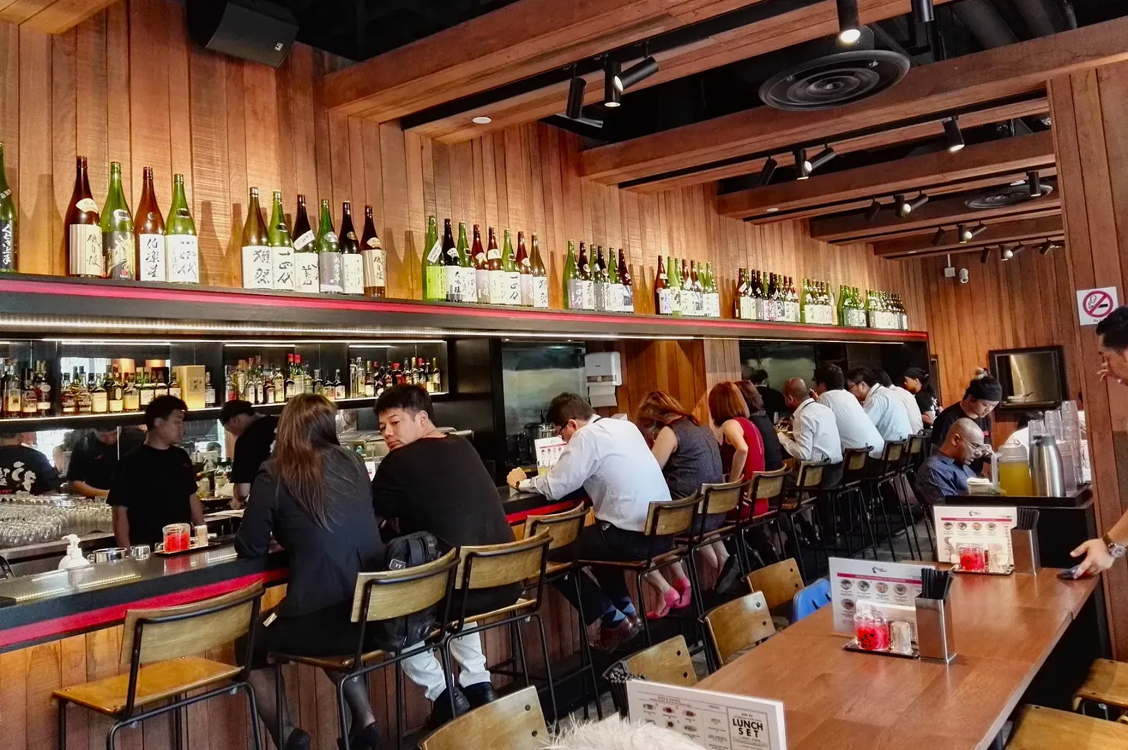A lively Japanese bar with patrons seated at a wooden counter under shelves of sake bottles. The warm lighting creates a cozy and inviting ambiance.
