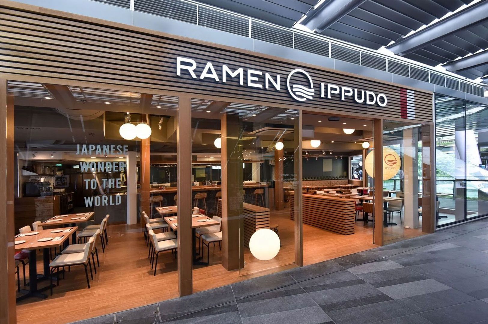 Exterior storefront view of a contemporary Ramen Ippudo restaurant with large glass windows, featuring the brand's logo and signage, and visible dining tables inside.