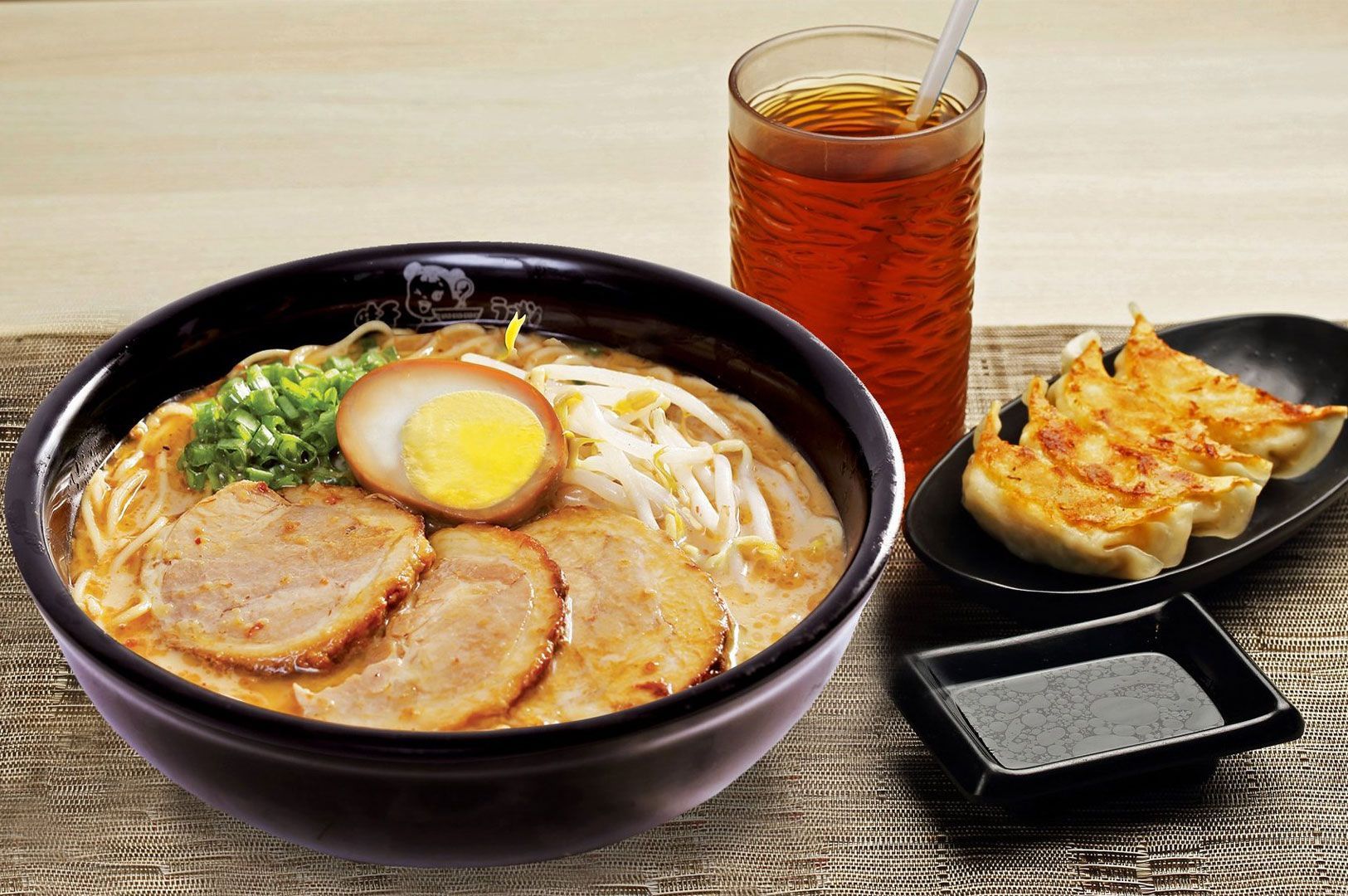 A complete meal set featuring a bowl of ramen topped with chashu pork and a seasoned egg, a side of pan-fried gyoza dumplings with dipping sauce, and a glass of iced tea.