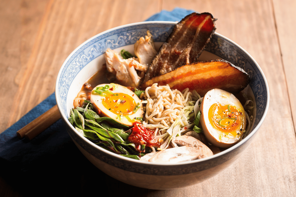 A visually striking bowl of gourmet ramen served in a blue-and-white patterned bowl on a wooden surface, featuring golden noodles, soft-boiled eggs with runny yolks, crispy-edged pork belly slices, fresh greens, mushrooms, green onions, and a touch of chili paste. A blue cloth napkin adds texture and warmth to the scene, highlighting the artisanal quality and ingredient complexity found in Singapore’s top-tier ramen offerings.