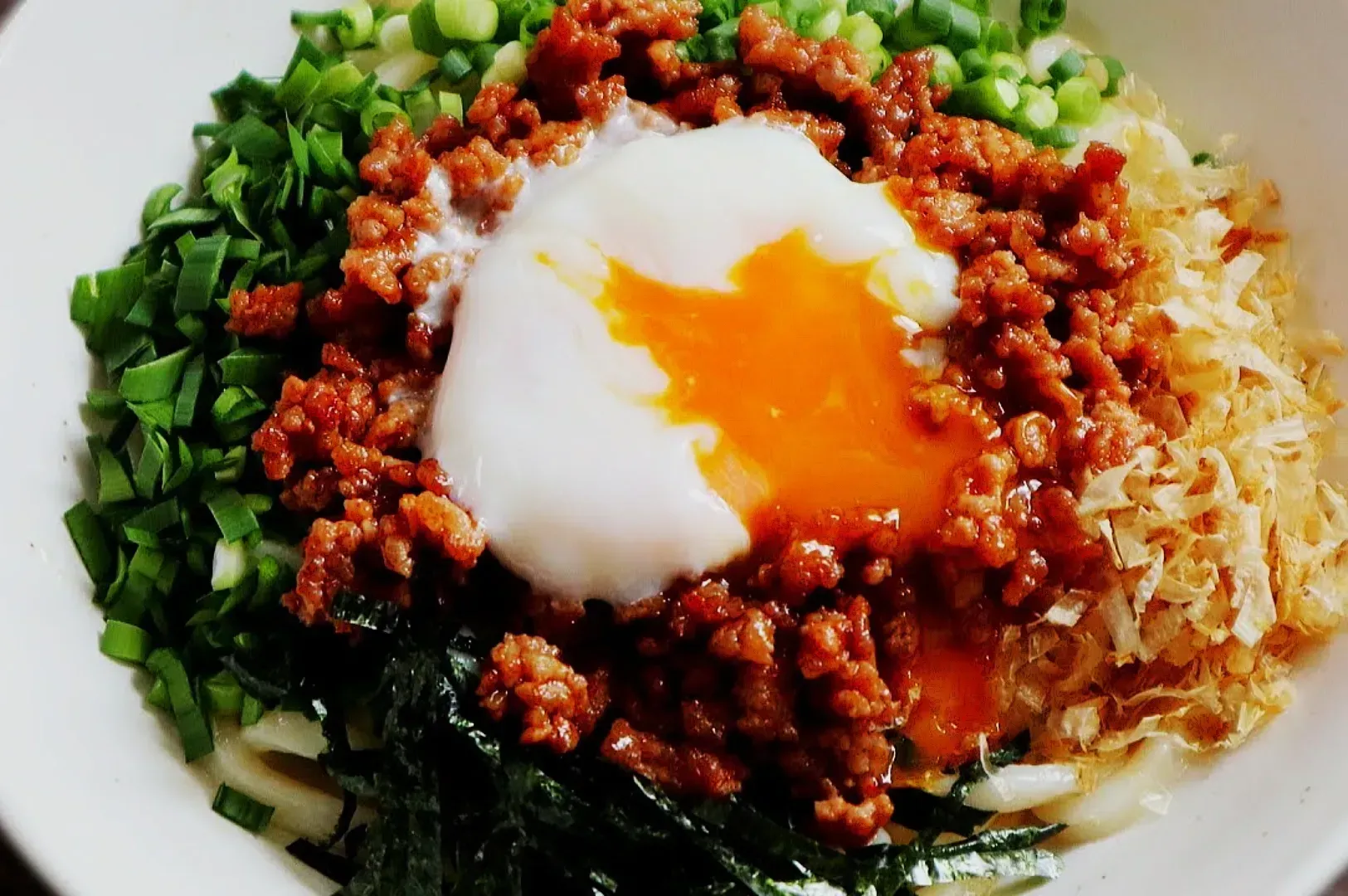A bowl of noodles topped with a perfectly poached egg, surrounded by minced meat, chopped green onions, shredded seaweed, and bonito flakes. The egg yolk is brightly golden, adding a rich and savory contrast to the vibrant green and brown ingredients.