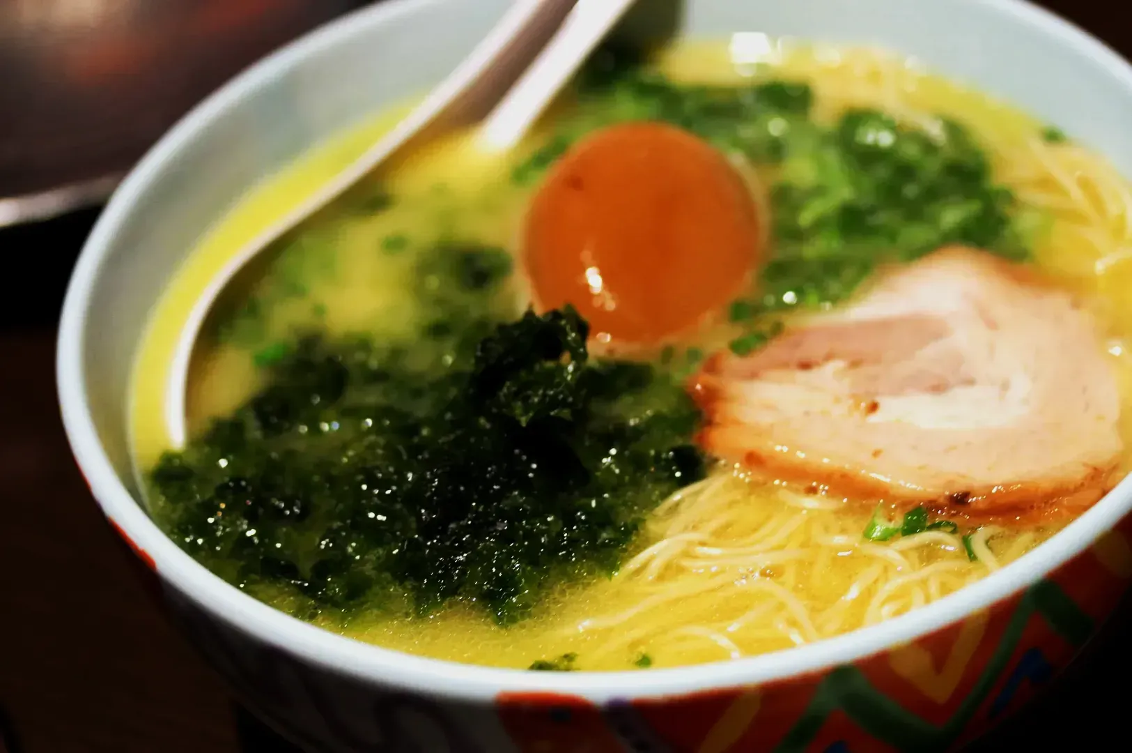 A steaming bowl of ramen with creamy broth, thin noodles, sliced pork, a marinated egg, and bright greens. The setting is warm and inviting.