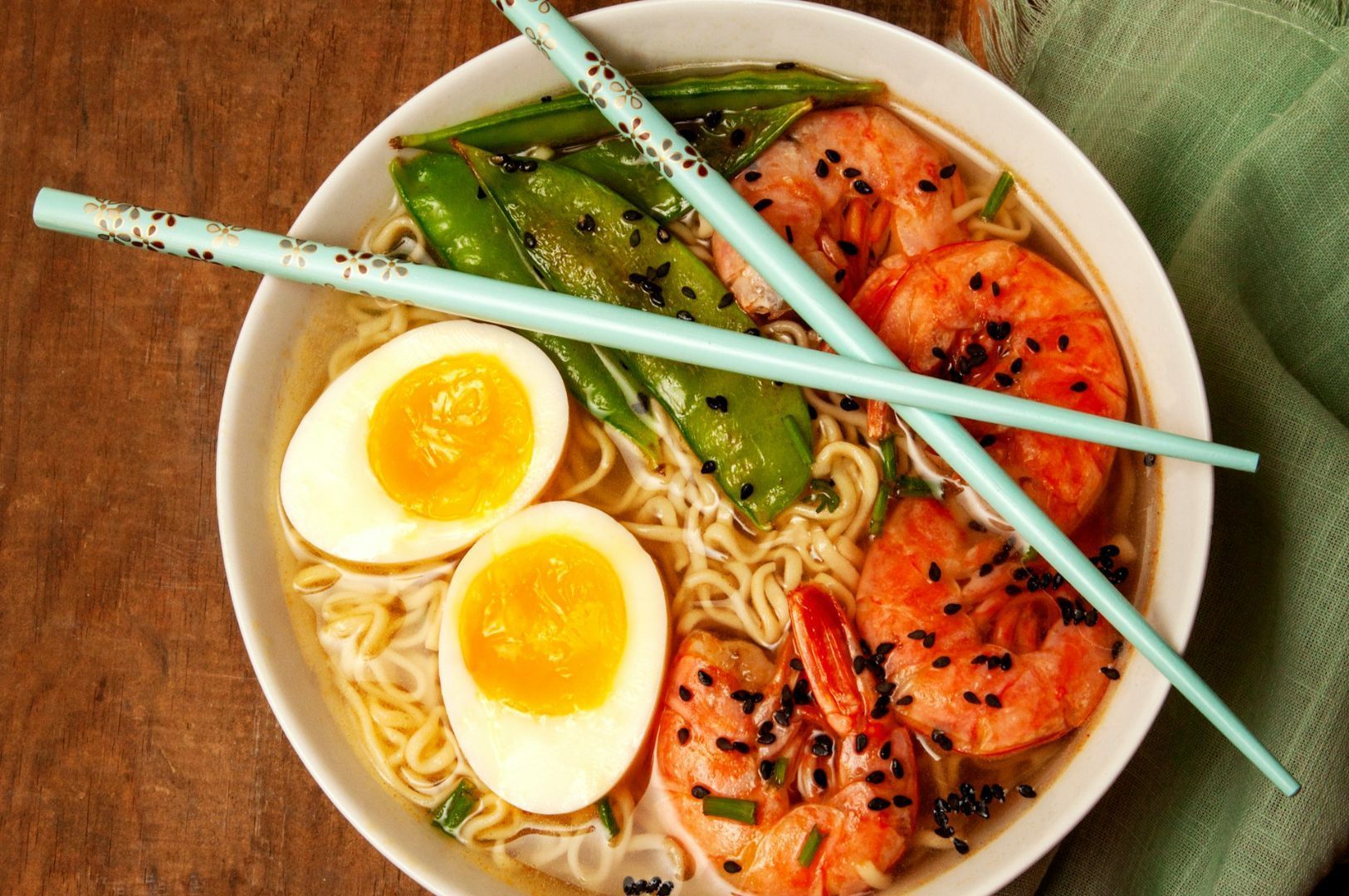 A colorful bowl of ramen with shrimp, snap peas, halved soft-boiled eggs, and black sesame seeds, topped with aqua chopsticks on a wooden table.