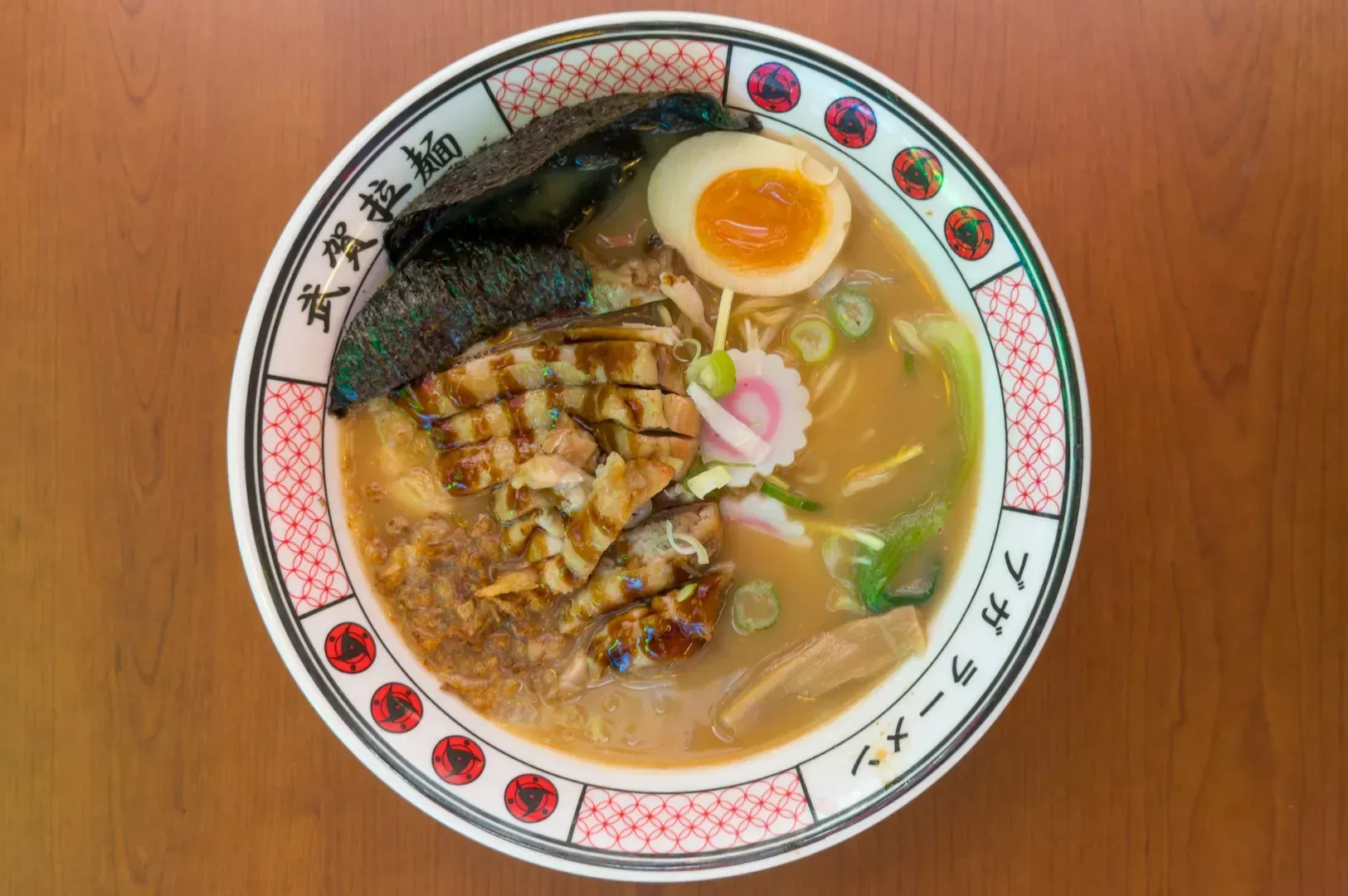 A bowl of ramen with grilled chicken, seaweed, soft-boiled egg, fish cake, and scallions in rich broth, served in a decorative bowl on a wooden table.