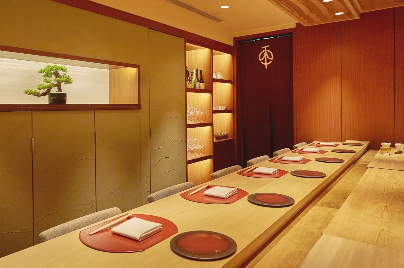Elegant Japanese dining room with a long wooden table set for eight, featuring red placemats and neatly folded napkins. A bonsai and shelves with glasses add a serene ambiance.