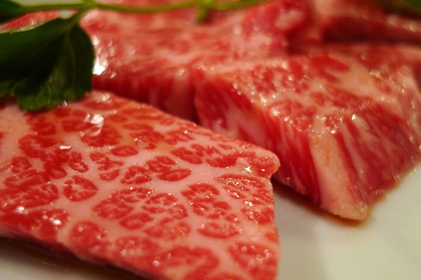 Close-up of marbled Wagyu beef slices on a plate, showcasing intricate fat patterns. A sprig of green parsley adds contrast. The tone is luxurious and appetizing.
