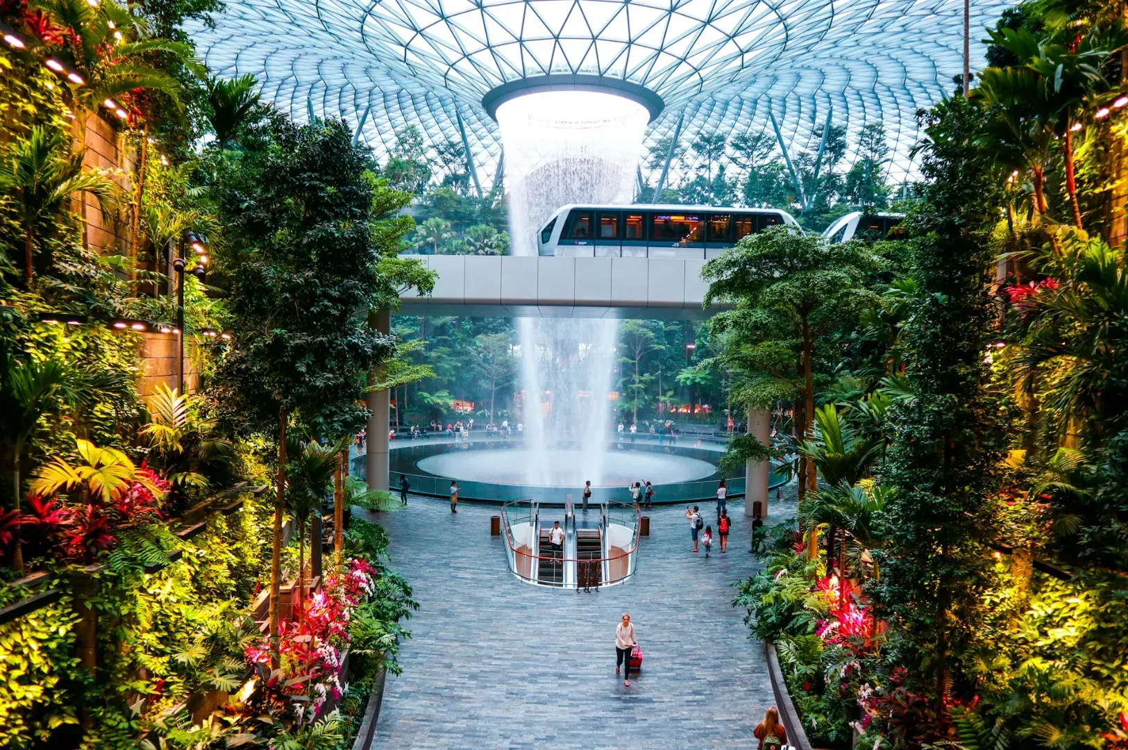 Lush indoor garden with cascading waterfall under a glass dome. A skytrain traverses above, and people stroll around, creating a serene urban oasis.