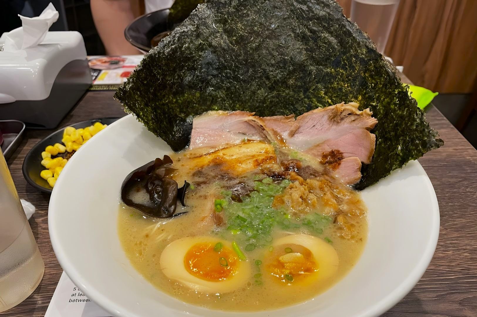 Bowl of creamy ramen with sliced pork, seaweed, soft-boiled eggs, and green onions. A small dish of corn and tissues are in the background.