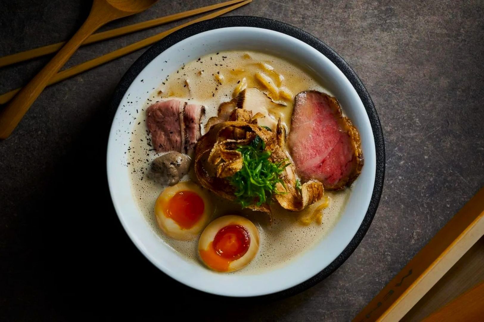 A bowl of creamy ramen with slices of rare beef, soft-boiled eggs, and scallions on a textured surface. Wooden utensils lie nearby, creating a cozy, gourmet vibe.