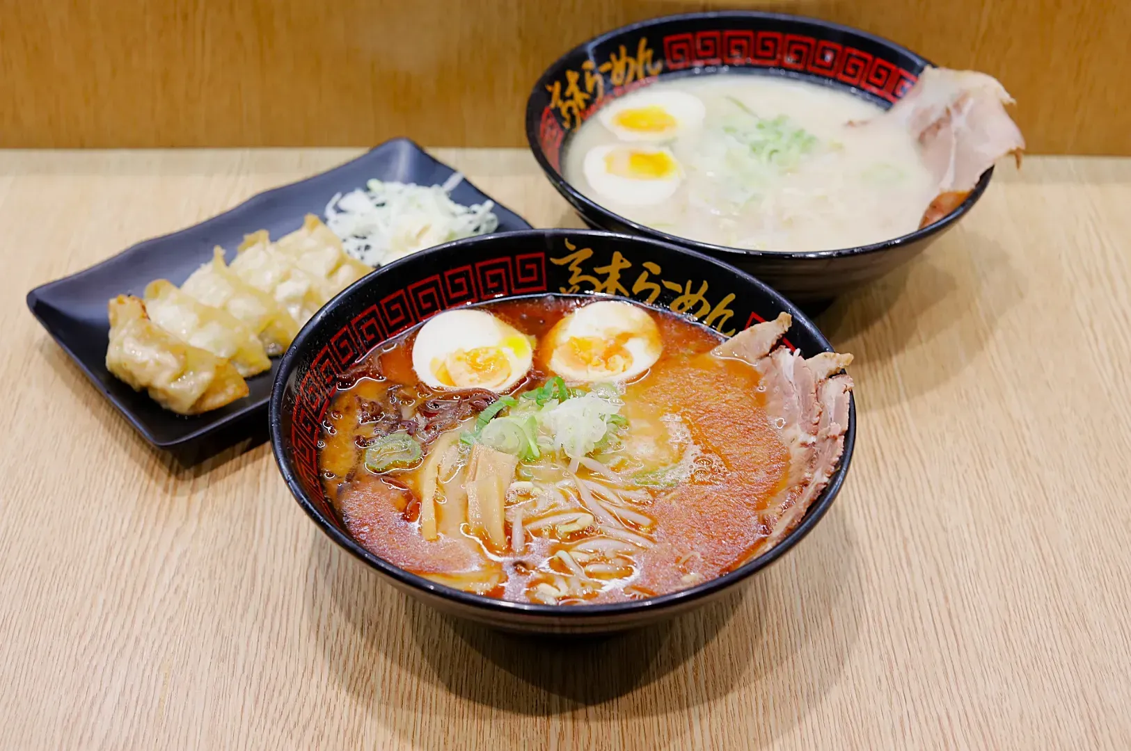 Two bowls of ramen sit on a wooden table. The front bowl has a rich brown broth, sliced pork, boiled eggs, and green onions. The rear bowl features a creamy broth with similar toppings. A black plate of golden gyoza dumplings with shredded cabbage is to the side. The scene conveys a warm, inviting atmosphere.